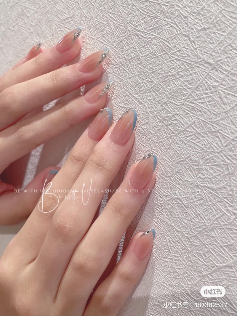Bàn tay với móng tay dài được trang trí sơn màu nude và xanh, đính đá, trên nền tường hoa văn trắng.