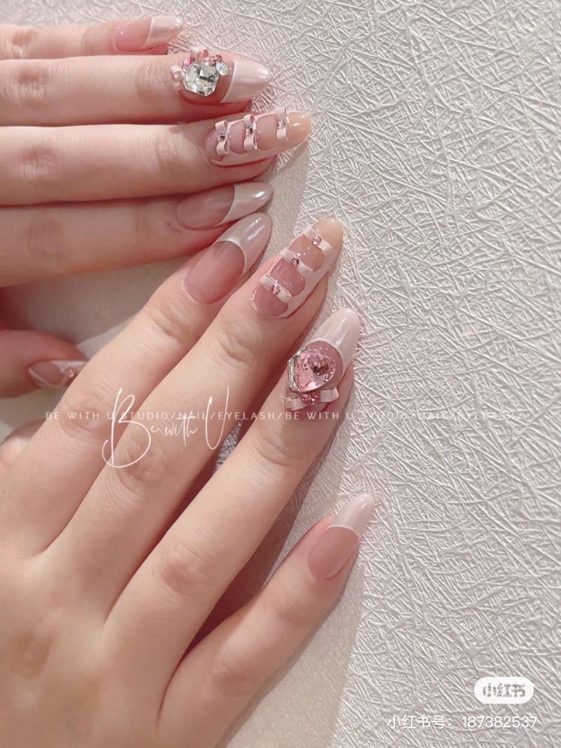 Bàn tay với móng tay trang trí họa tiết và đính đá, nền tường màu sáng có hoa văn.