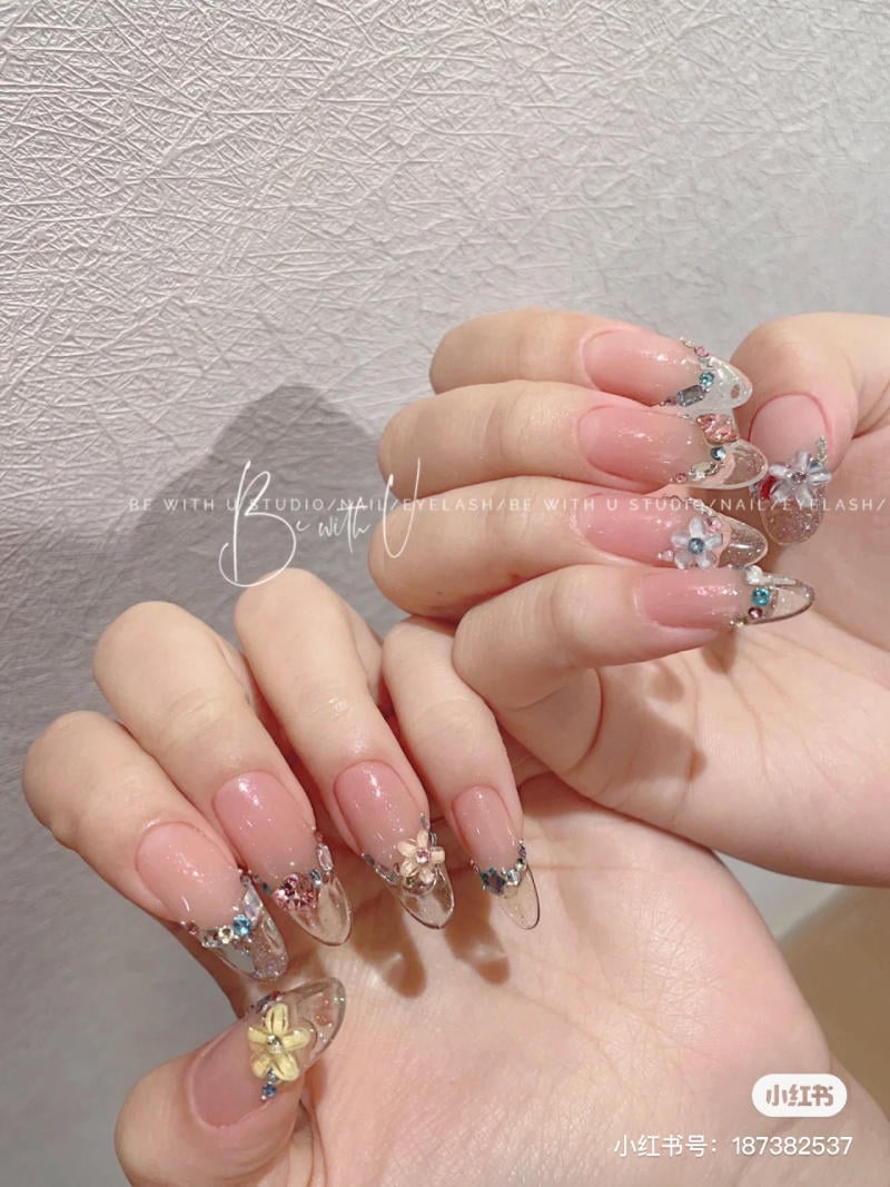 Bàn tay với móng tay dài, được trang trí hoa và đá lấp lánh, nền tường màu xám nhạt.