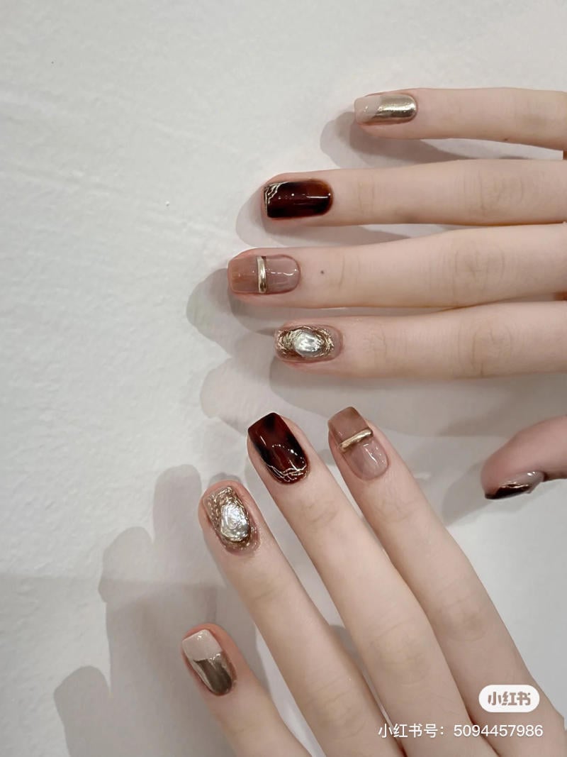Bàn tay với móng tay được sơn và trang trí họa tiết màu nâu và vàng trên nền trắng.