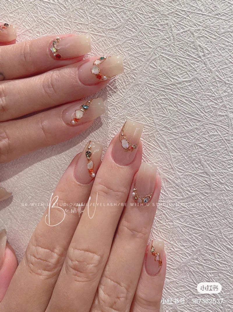 Bàn tay với móng tay được trang trí bằng đá màu và họa tiết ánh kim trên nền móng màu nude, trên nền vải trắng.