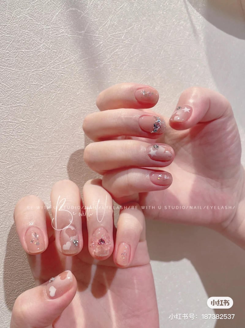 Bàn tay với móng tay được trang trí họa tiết lấp lánh và hình trái tim trên nền màu hồng nhạt.