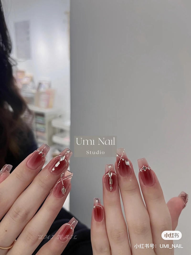 Bàn tay với móng tay được trang trí màu đỏ ombre và đính hạt lấp lánh, nền là không gian tiệm nail.