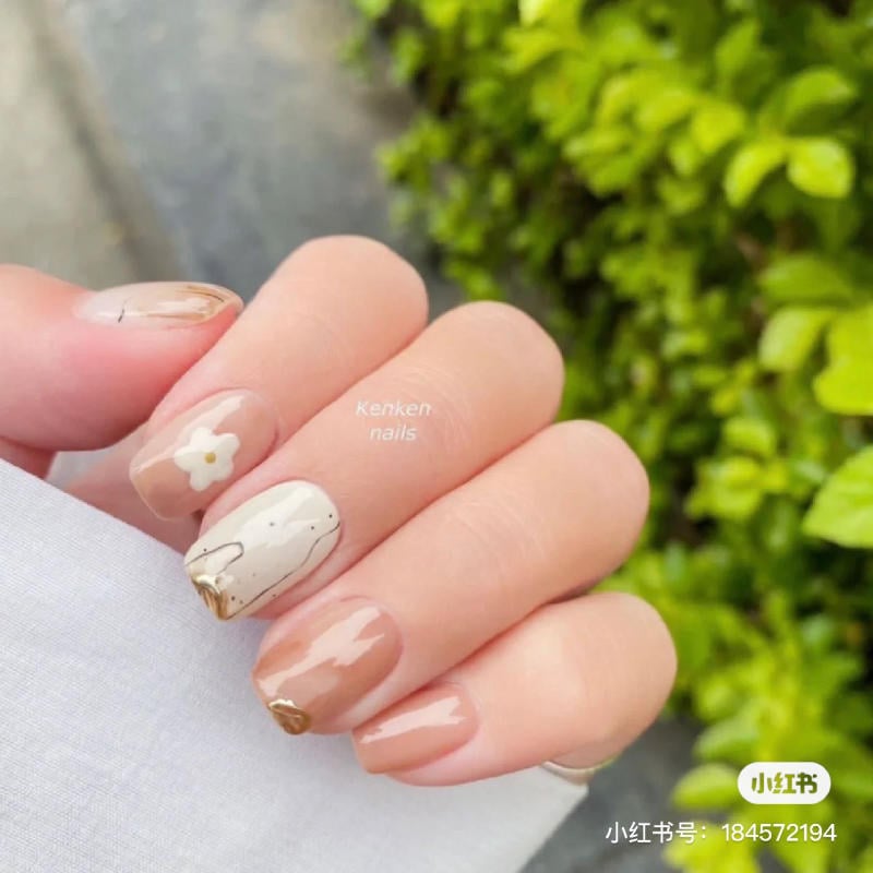 Bàn tay với móng tay được sơn màu nude và trắng, có họa tiết hoa và đường vẽ trang trí, nền lá cây xanh.