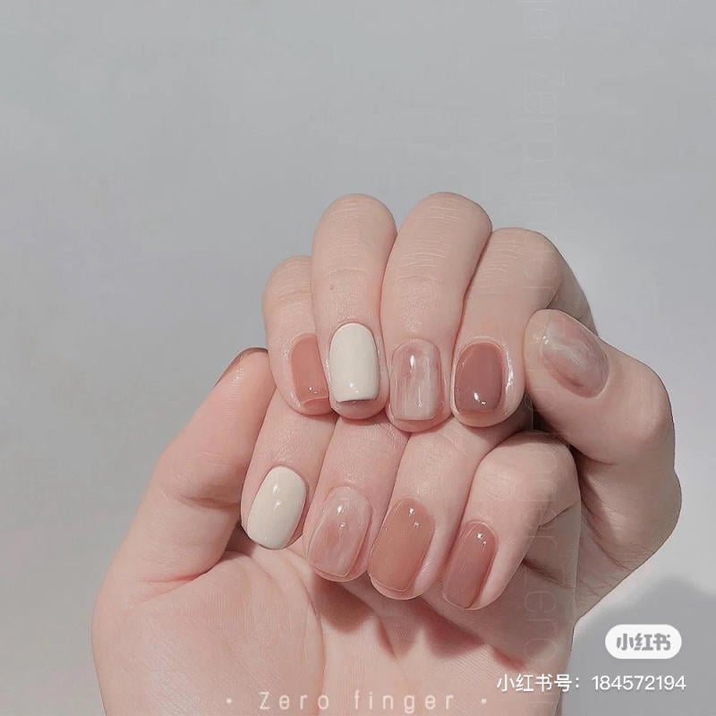 Bàn tay với móng tay được sơn màu nude và trắng, trên nền xám nhạt.