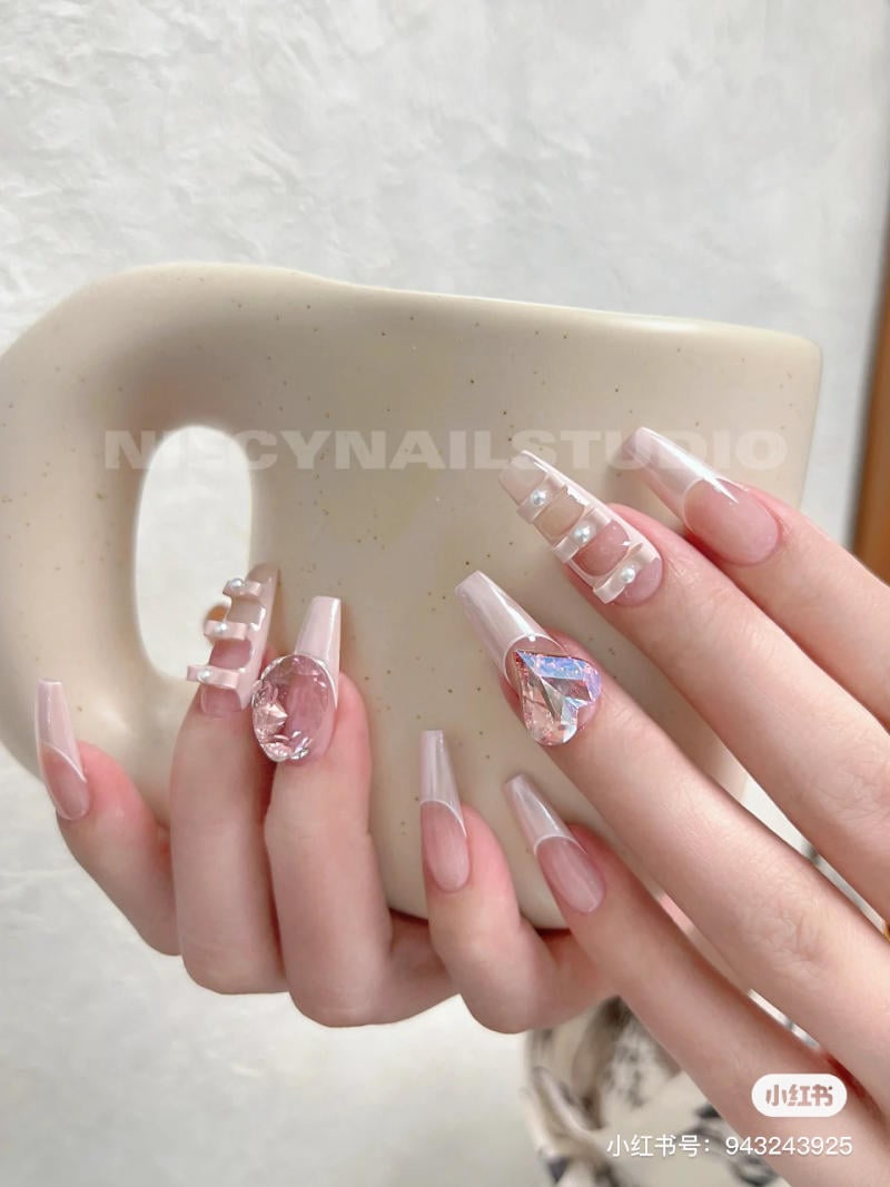 Bàn tay với móng tay dài, được trang trí bằng đá quý và ngọc trai, đang cầm một chiếc cốc màu kem.