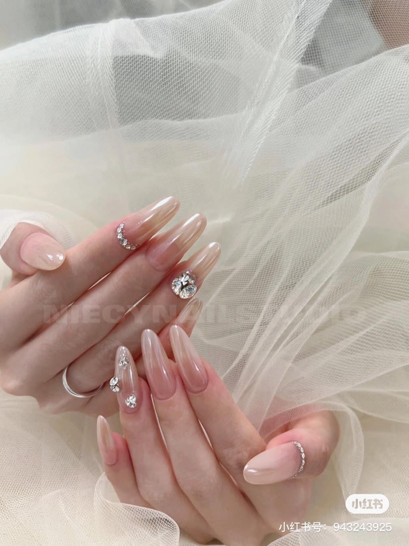 Bàn tay với móng tay dài, sơn màu nude và đính đá, trên nền vải trắng mỏng.