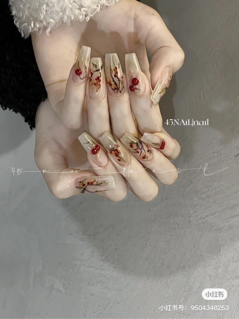 Bàn tay với móng tay dài, trang trí hoa văn và trái cây màu đỏ trên nền nhạt, cầm nhẹ nhàng trước nền xám.