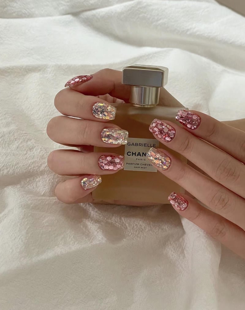 mau-nail-nhu-dep__26__2a1d4f7a32a94862bcd499c091deb692 Bàn tay với móng tay đính đá lấp lánh cầm chai nước hoa nằm trên nền vải trắng.