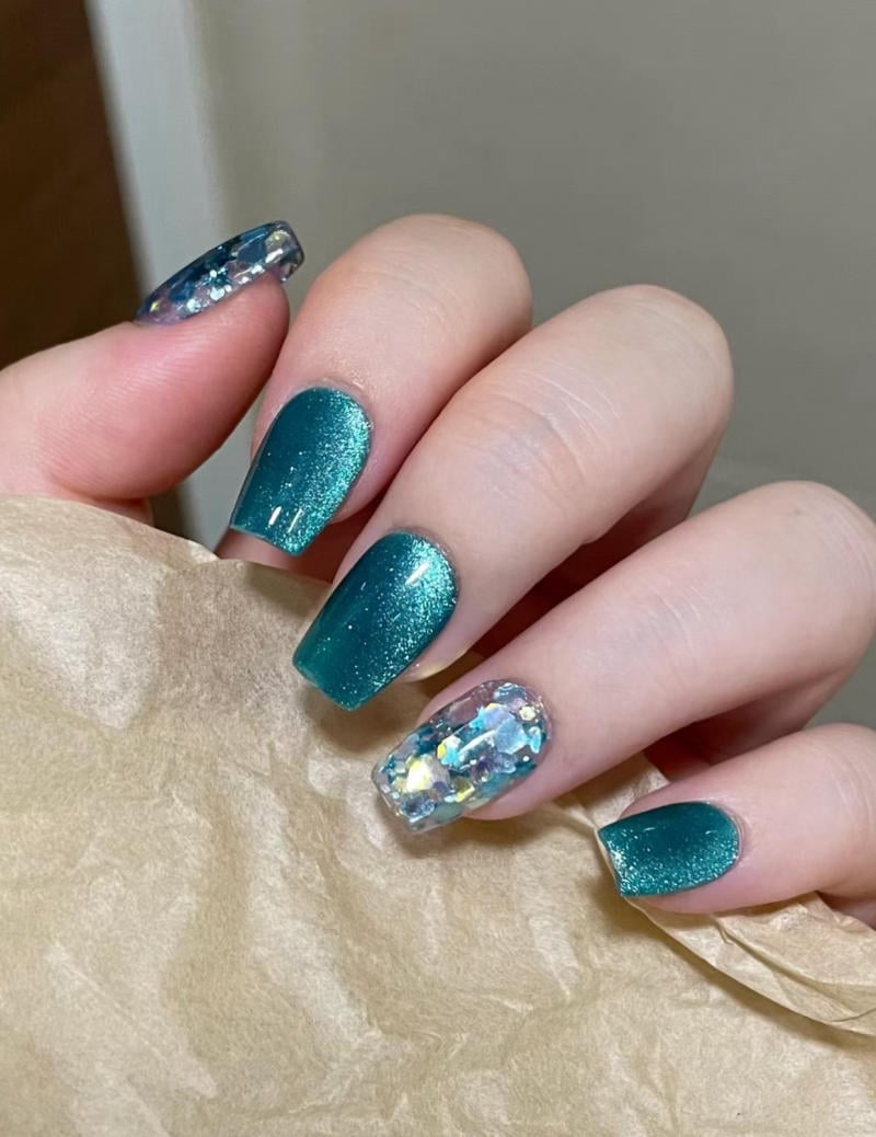 mau-nail-nhu-dep__27__8ff190c6948d4455a299c01c7c7ba720 Bàn tay với móng tay sơn màu xanh lấp lánh và một móng đính đá, cầm một tờ giấy màu nâu.