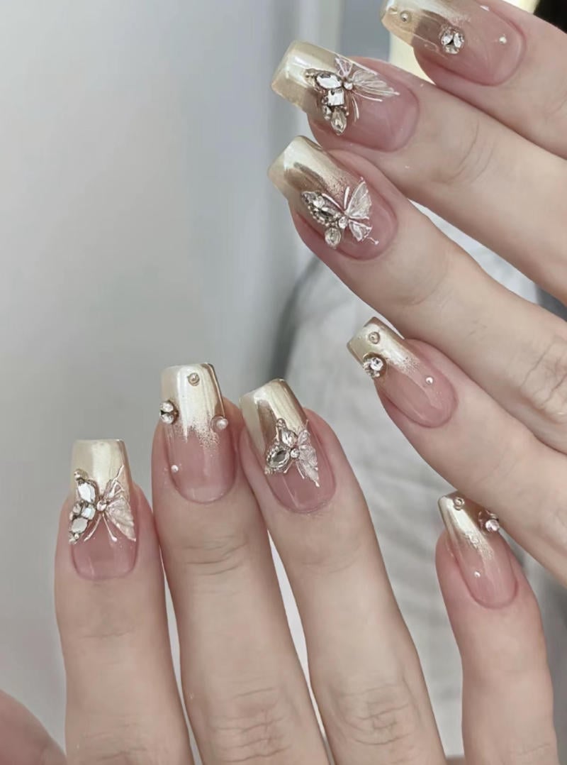 mau-nail-nhu-dep__30__6b1772b909764023a9cd411ae0280683 Bàn tay với móng tay được trang trí màu vàng nhạt, đính đá và hình bướm lấp lánh.