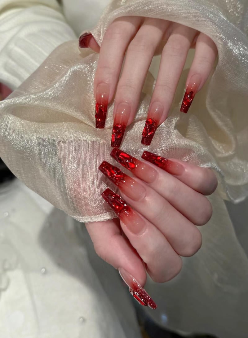 mau-nail-nhu-dep__40__6102c076541b4e14bd67c2ab18e0b0f1 Bàn tay với móng tay dài, được trang trí họa tiết đỏ lấp lánh, đang cầm một tấm vải mỏng màu trắng.