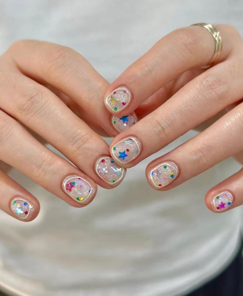 mau-nail-nhu-dep__42__23bd227ad76c409797c36261d2b56e31 Bàn tay với móng tay được sơn màu nhũ, trang trí các hình ngôi sao và chấm tròn nhiều màu sắc.
