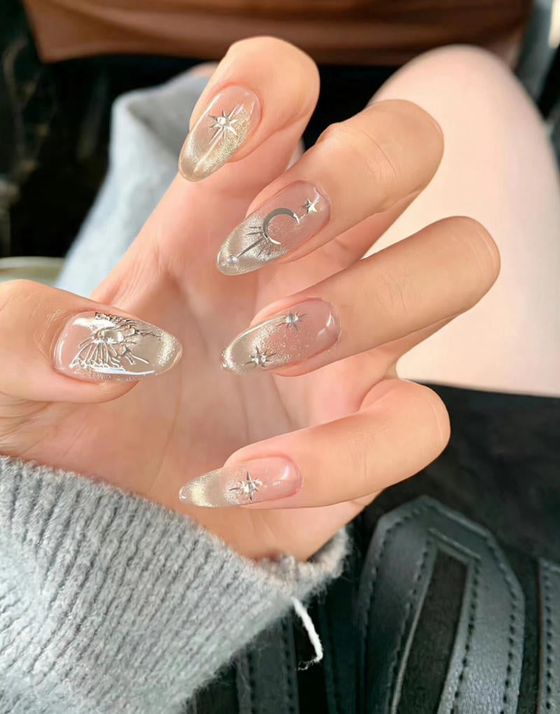 mau-nail-nhu-dep__46__00142a5cdd464cca95ec2aae5080b9ae Bàn tay với móng tay trang trí họa tiết kim loại hình ngôi sao và mặt trăng trên nền màu nhạt.