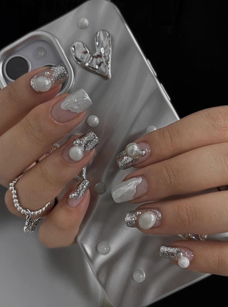 mau-nail-nhu-dep__47__3eb95be023cc4597aed2ad1c4ca6a675 Bàn tay với móng tay được trang trí họa tiết bạc và ngọc trai, cầm điện thoại có ốp lưng màu bạc.