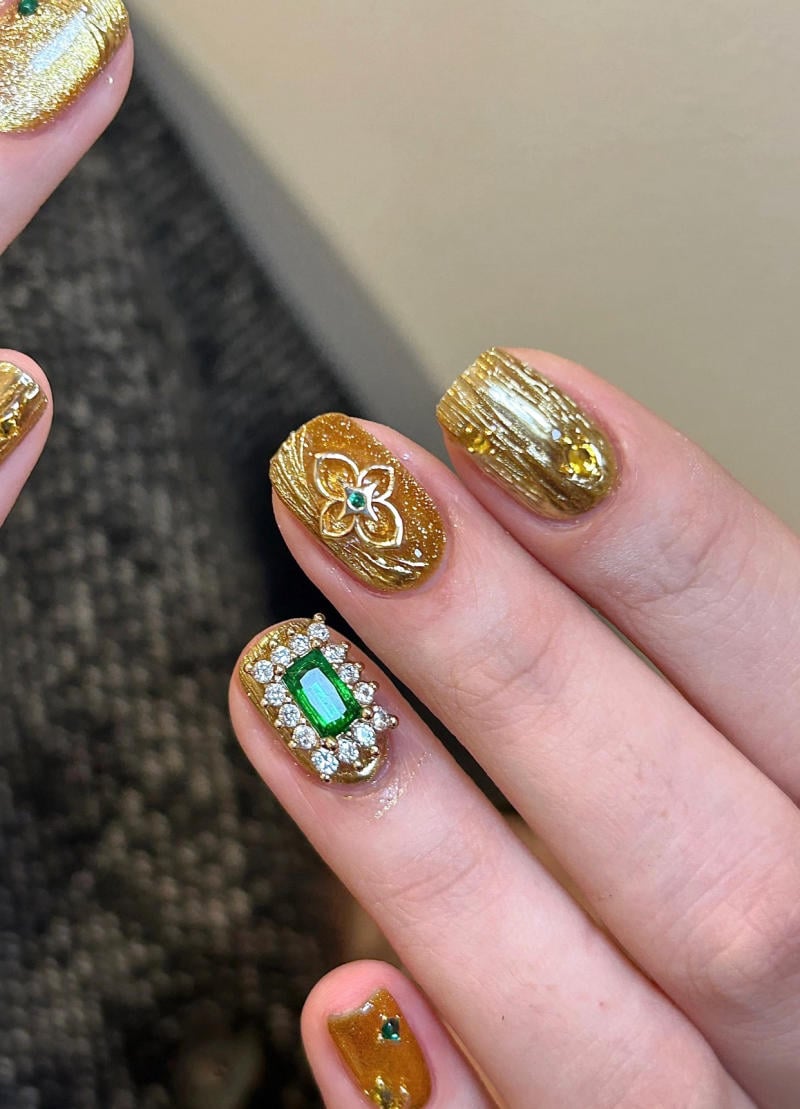 mau-nail-nhu-dep__53__b81d695069ef4a09b7a06143874ac7ee Bàn tay với móng tay màu vàng ánh kim, được trang trí hoa văn và đính đá lấp lánh.