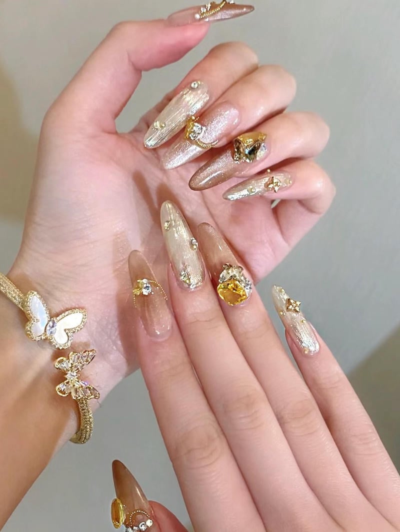mau-nail-nhu-dep__56__cc6c291286d84704ba80a334115b6862 Bàn tay với móng tay dài được trang trí lấp lánh và phụ kiện hình bướm, nền màu trung tính.