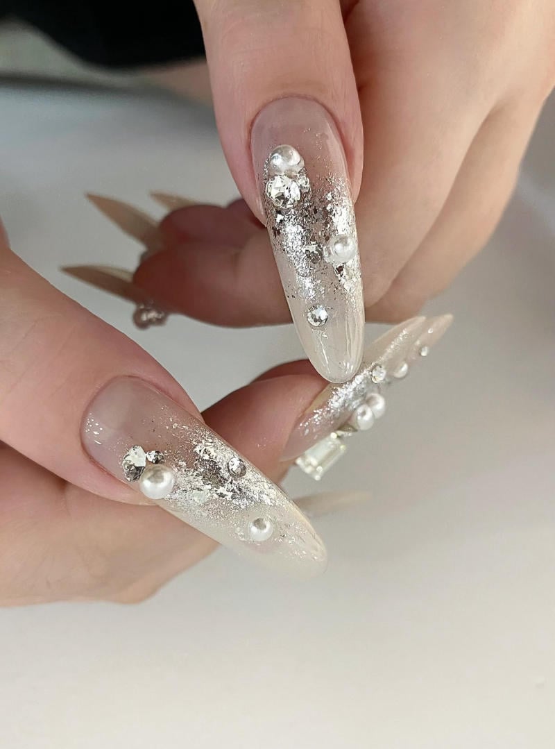 mau-nail-nhu-dep__59__2be65953e6034939bd796228dc488270 Bàn tay với móng tay dài được trang trí bằng họa tiết ngọc trai và kim loại lấp lánh trên nền màu sáng.