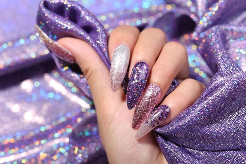 mau-nail-nhu-dep__6__570b019c47df4ab6b90eaaaa1fb77c19 Bàn tay với móng tay lấp lánh nhiều màu sắc, đang nắm vải ánh kim màu tím.