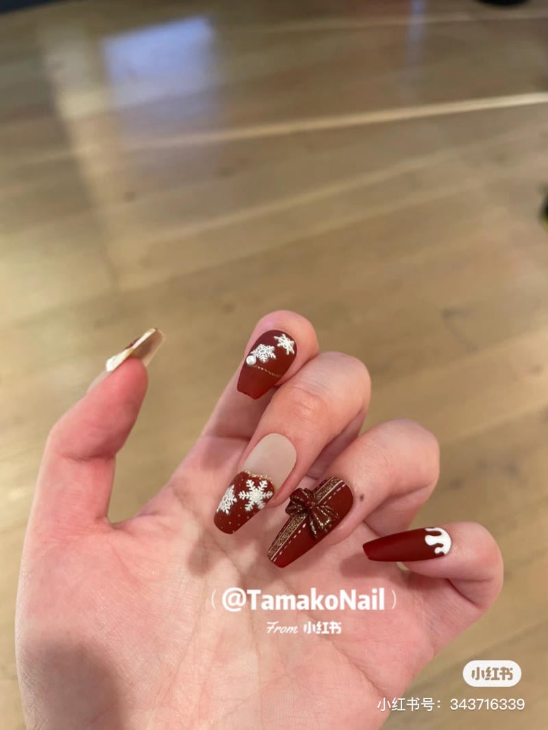 mau-nail-noel-dep__10__b57171260f5d44d1afb6799789693e58 Bàn tay với móng tay được sơn màu đỏ, trang trí họa tiết hoa tuyết và nơ, trên nền sàn gỗ.