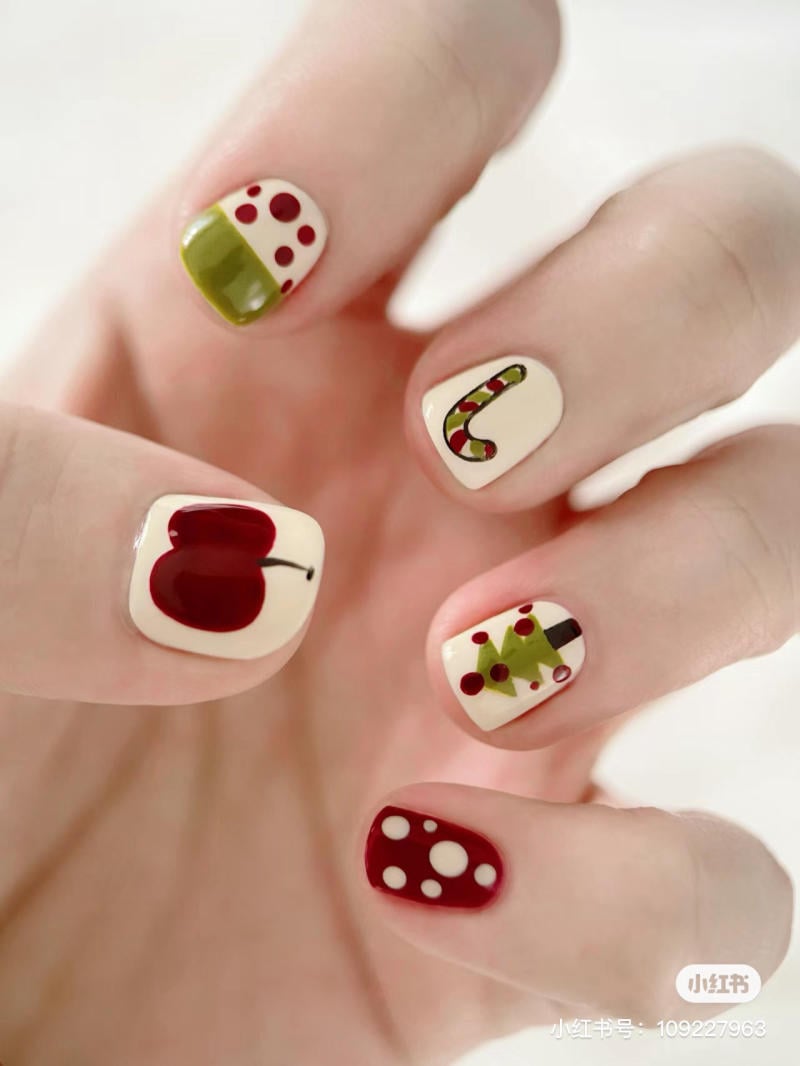 mau-nail-noel-dep__14__8f4fbef4d0cd495bacfb2e41057494da Bàn tay với móng tay được trang trí họa tiết trái cây, cây thông và chấm bi màu đỏ, xanh lá trên nền trắng.