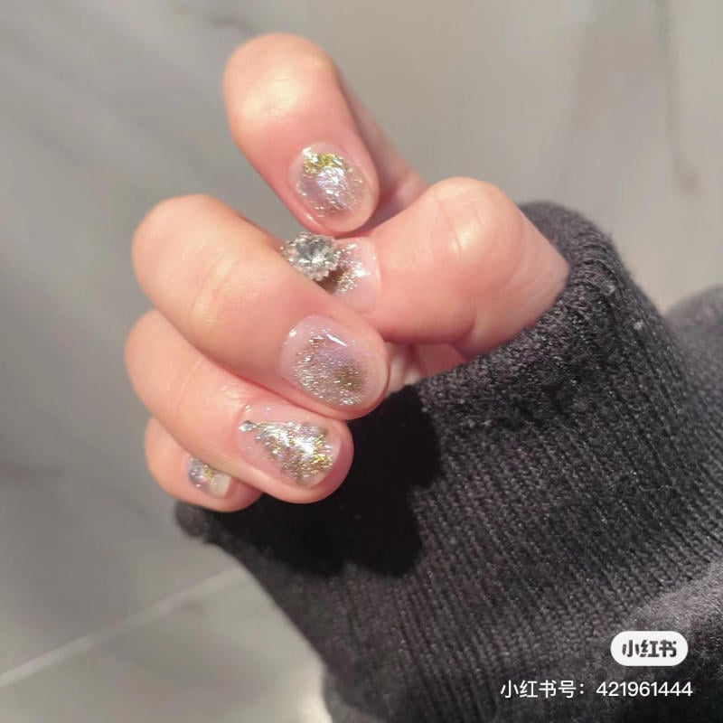 mau-nail-noel-dep__46__7d9ac6c39e8246bab835b7a1f5e9faba Bàn tay với móng tay được sơn màu lấp lánh, mặc áo len đen, trong không gian nền xám.