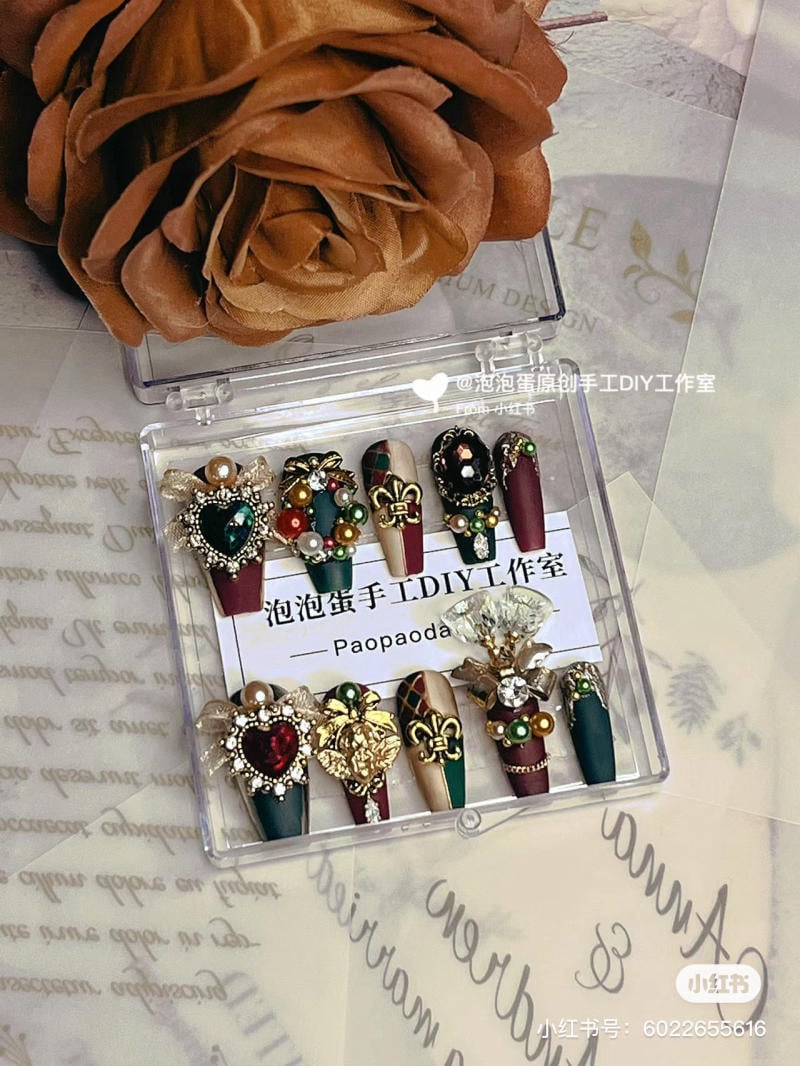 mau-nail-noel-dep__48__03165fdaa44243f892e0d3aaf8c7db9a Hộp móng giả trang trí lộng lẫy với họa tiết và đá quý, nằm cạnh hoa hồng màu nâu.