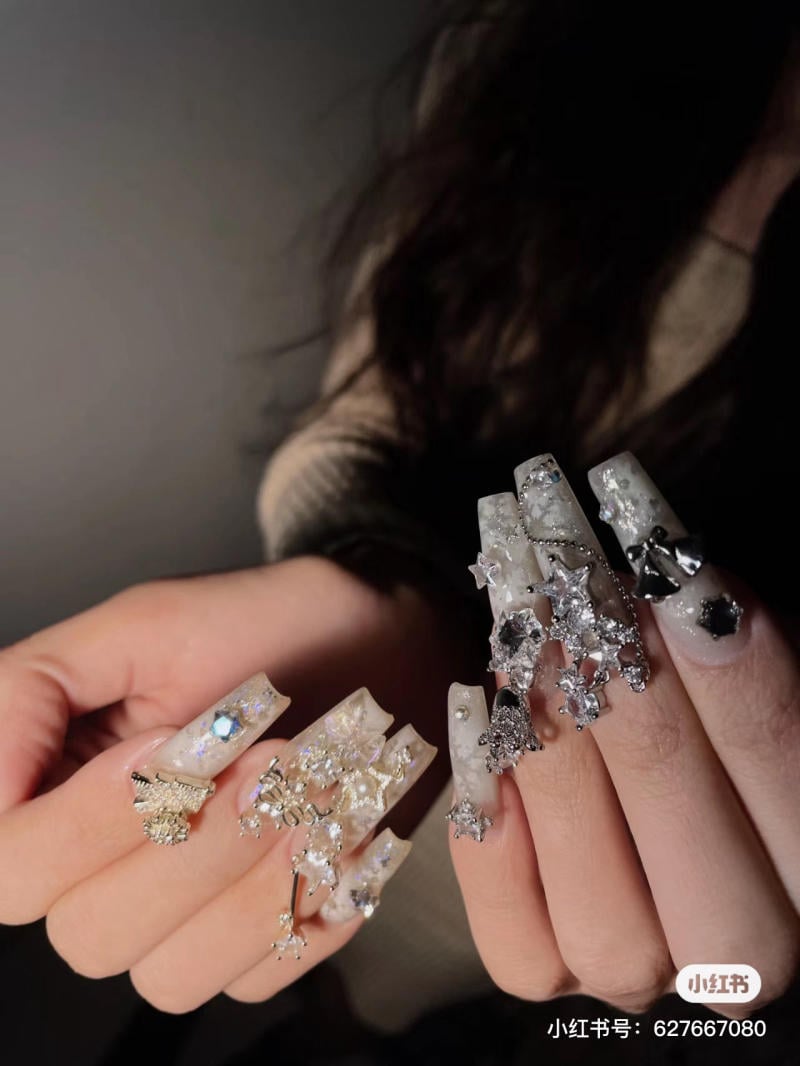 mau-nail-noel-dep__4__fa6faf564ebf486a95c7c2b88d2f82aa Bàn tay với móng tay dài, được trang trí bằng hoa văn và phụ kiện lấp lánh, nổi bật dưới ánh sáng.