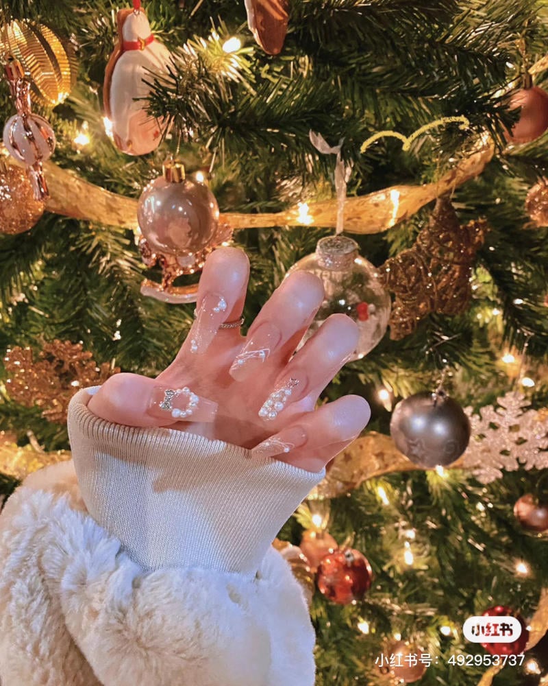 mau-nail-noel-dep__51__5ae7b61ad6134b3fafd155f5894e3a16 Bàn tay với móng tay trang trí đứng trước cây thông Noel có đèn và đồ trang trí lấp lánh.