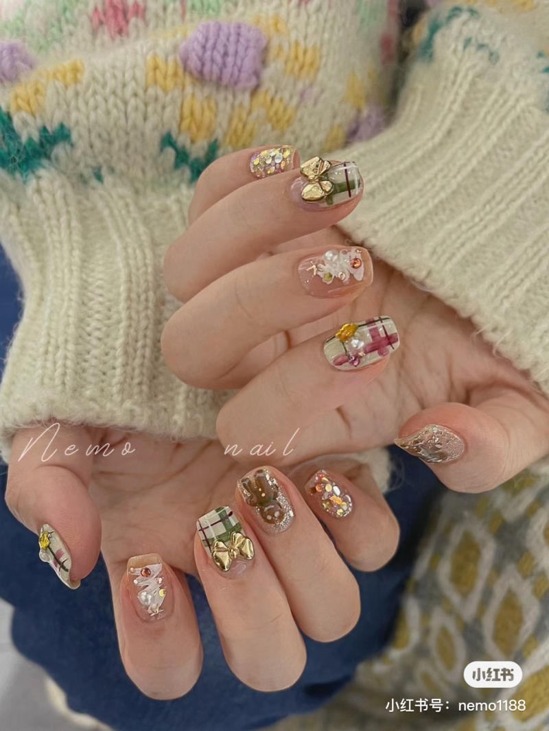 mau-nail-noel-dep__5__656c7ac156d149b3b0b1ee314fa20681 Bàn tay với móng tay được trang trí họa tiết màu sắc và phụ kiện nổi bật, mặc áo len dày màu sáng.