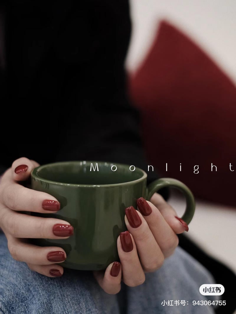 mau-nail-noel-dep__62__51ea95515c7644d8adde88dfc8b5c5e5 Người cầm ly màu xanh lá cây, móng tay sơn đỏ, nền có gối màu đỏ.