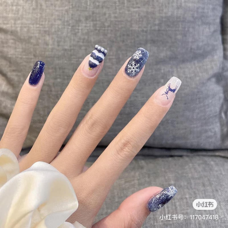 mau-nail-noel-dep__64__2143535adeba481b83718edc50411ded Bàn tay với móng tay được trang trí họa tiết mùa đông, màu xanh đậm và trắng, có hình bông tuyết và tuần lộc.