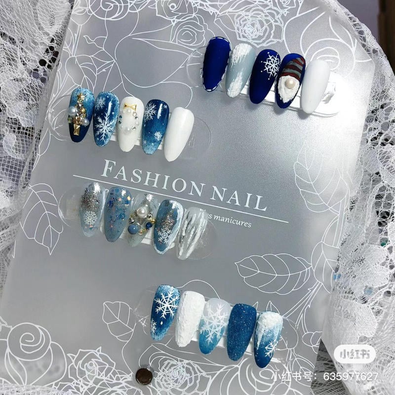 mau-nail-noel-dep__74__04328683dd304380941294bbc06cf92e Bộ móng giả với họa tiết tuyết và màu sắc xanh, trắng, được trưng bày trên nền giấy có hoa văn.