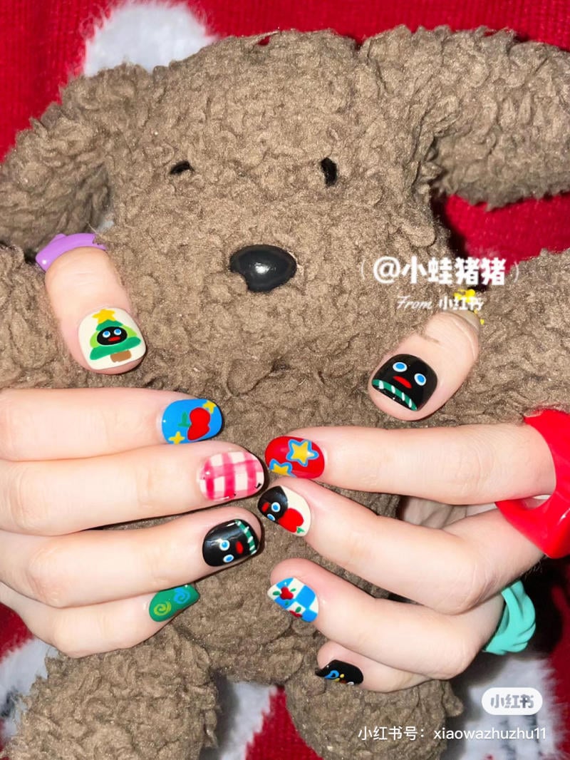 mau-nail-noel-dep__78__36528b6ef6c443cf92204ce8d3b7dc66 Bàn tay với móng tay vẽ họa tiết nhiều màu sắc đang ôm một chú gấu bông màu nâu.