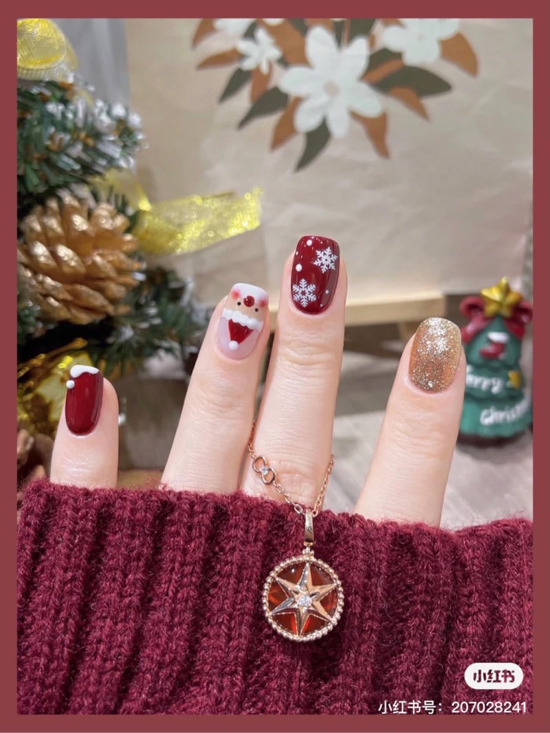 mau-nail-noel-dep__81__e149f3aff35b4d78bdf4d05e8d7098d3 Bàn tay với móng tay trang trí họa tiết Giáng sinh, đeo vòng cổ, nền cây thông Noel và hoa trắng.