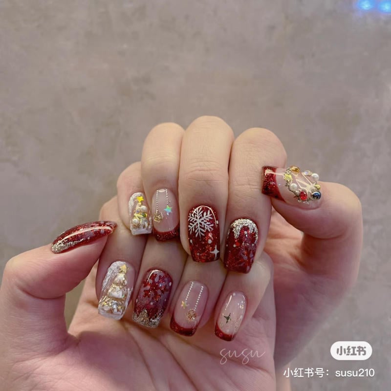 mau-nail-noel-dep__88__9988ed9ea5c5419ebb8eff79cb91e24d Bàn tay với móng tay trang trí họa tiết Giáng Sinh, nền đỏ, các hình trang trí như bông tuyết và cây thông.