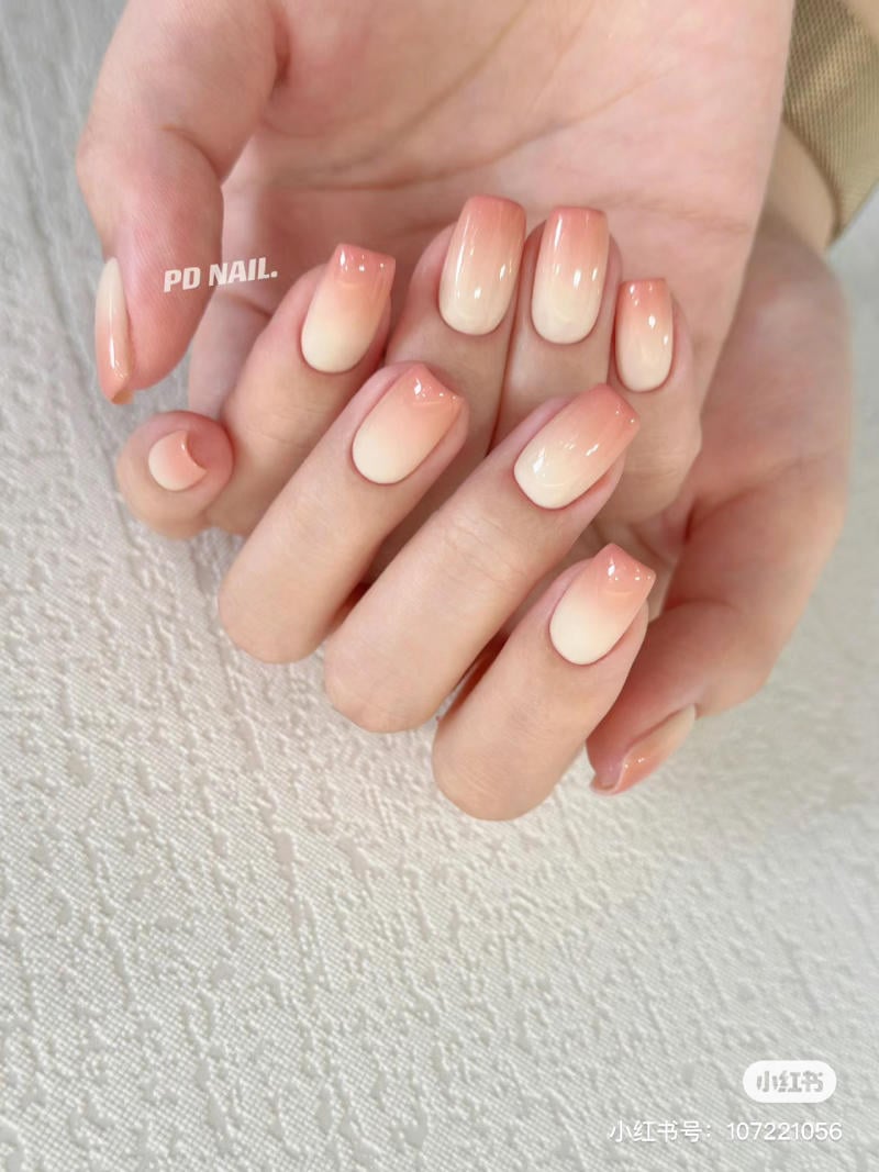 Bàn tay với móng tay được sơn màu ombre hồng nhạt, đặt trên nền vải trắng có họa tiết.