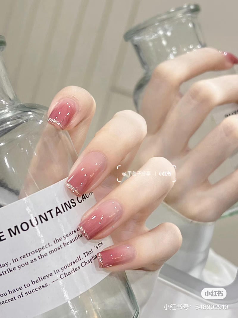 Bàn tay với móng tay sơn màu hồng nhạt cầm chai thủy tinh trong suốt, phản chiếu trong gương.