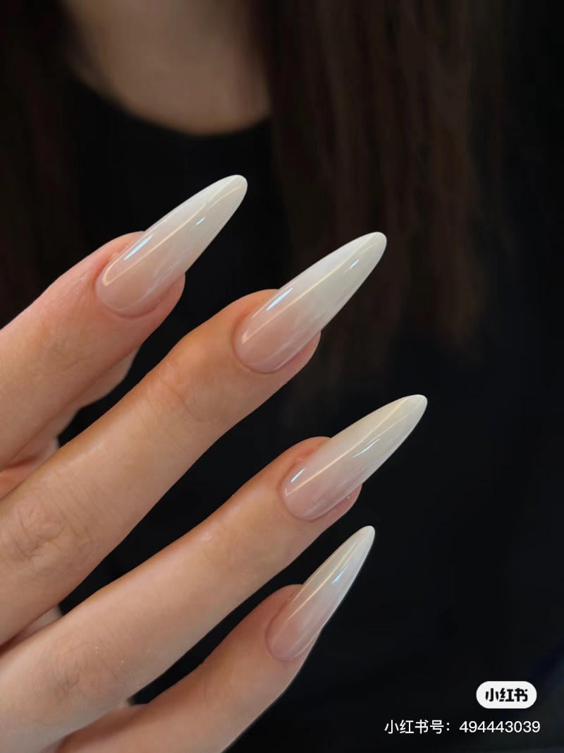 Bàn tay với móng tay dài, màu trắng trong suốt, tạo dáng nhọn, trên nền tối.