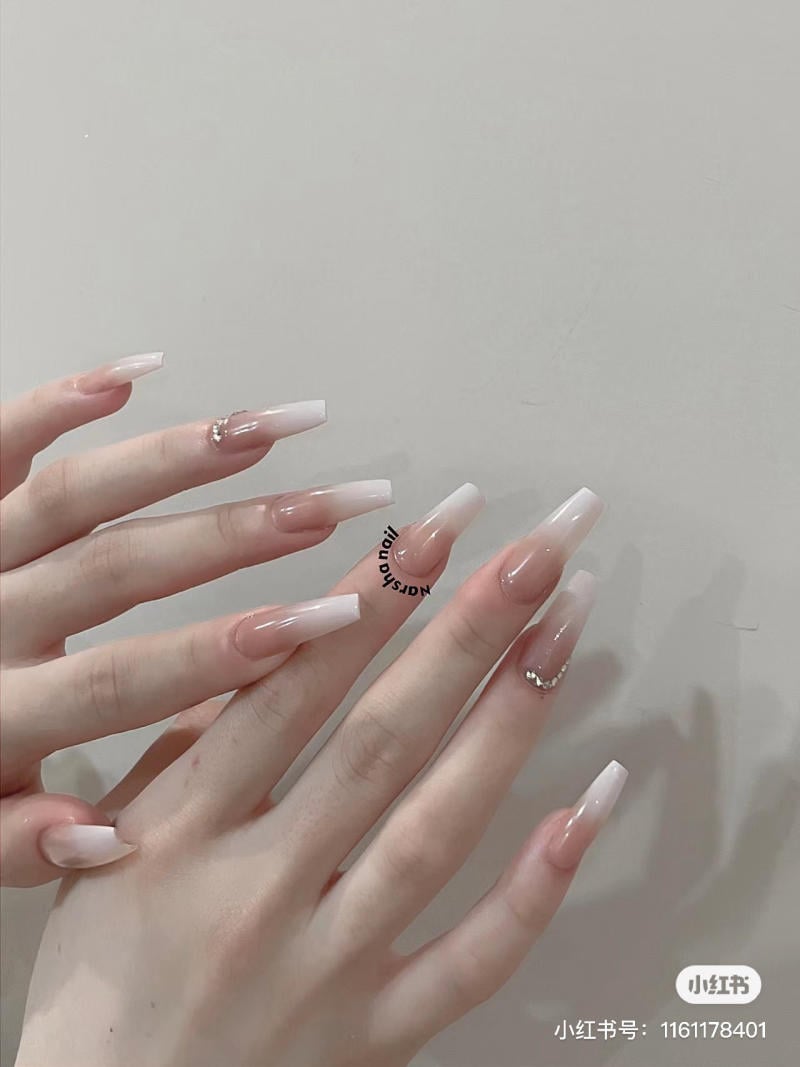 Bàn tay với móng tay dài, sơn màu hồng nhạt và đính đá, trên nền xám nhạt.