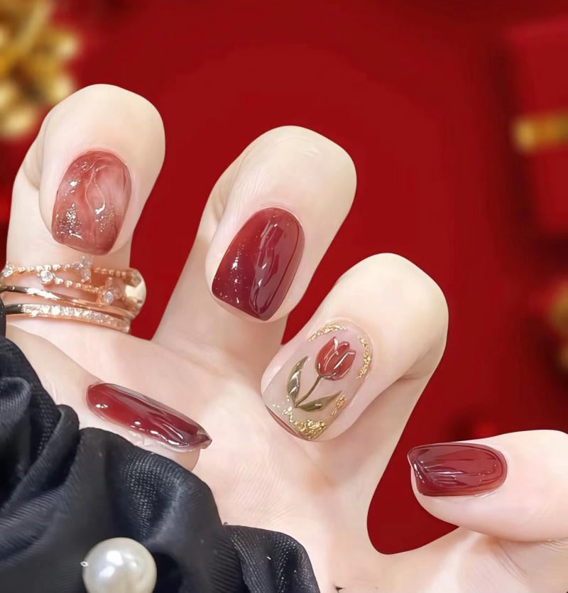Bàn tay với móng tay sơn màu đỏ, một móng có họa tiết hoa, nền đỏ, có nhẫn và khăn đen đính ngọc trai.