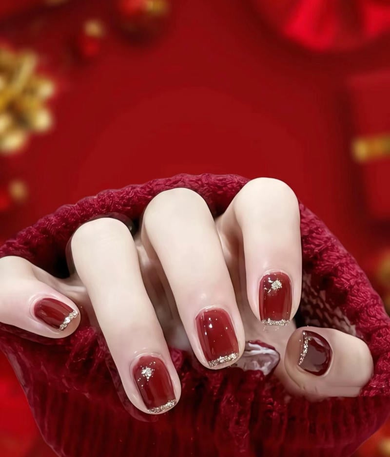 Bàn tay với móng tay sơn đỏ, viền ánh kim, cầm áo len đỏ, nền đỏ phía sau.