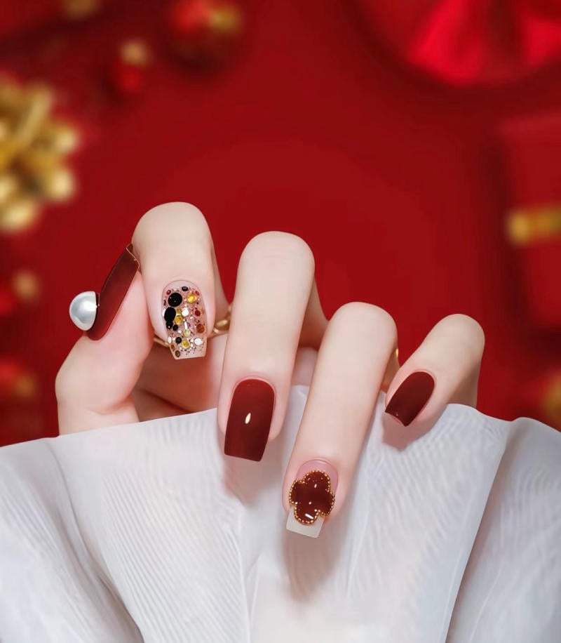 Bàn tay với móng tay được trang trí công phu, sơn màu đỏ và đính đá, nền đỏ và chi tiết vàng mờ.