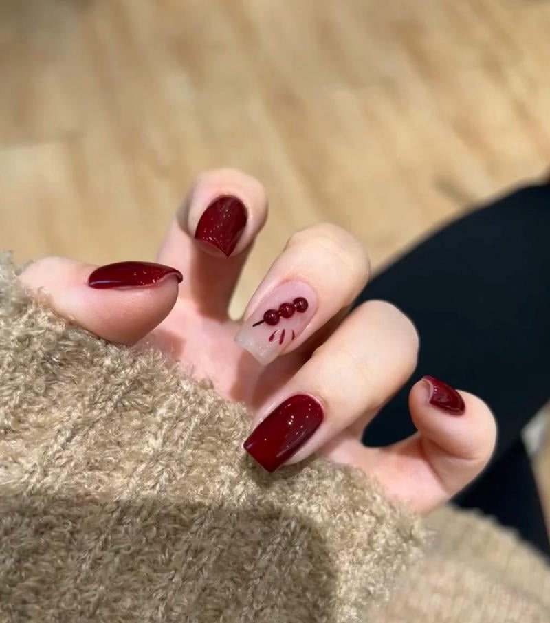 Bàn tay với móng tay sơn màu đỏ đậm, một móng có họa tiết trang trí nổi bật trên nền màu sáng.