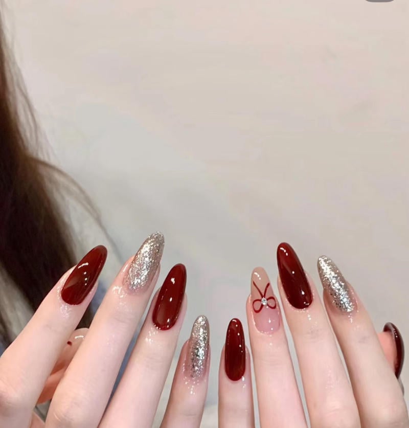 Bàn tay với móng tay dài, sơn đỏ và nhũ bạc, có hình nơ trên ngón áp út, nền trắng.