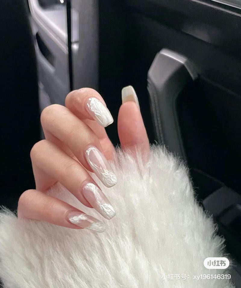 Bàn tay với móng dài, sơn màu trắng vân nổi, đặt trên áo lông trắng, trong xe ô tô.