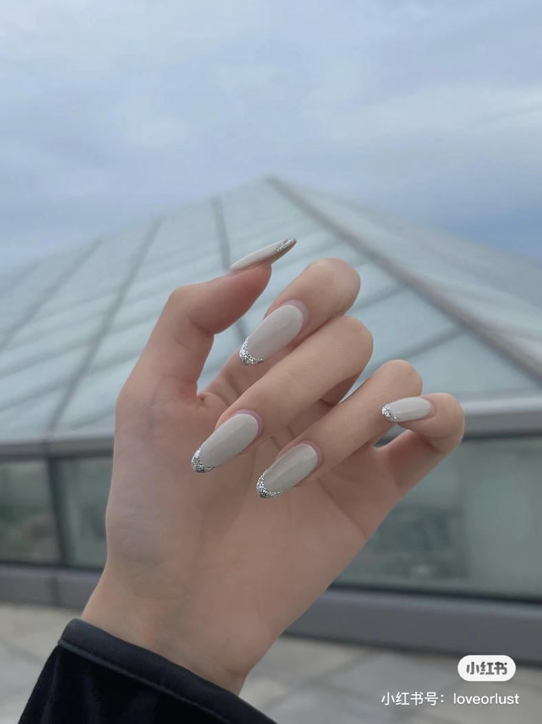 Một bàn tay với móng tay dài, sơn màu nude và đầu móng đính kim tuyến, nền là bầu trời và mái nhà kính.