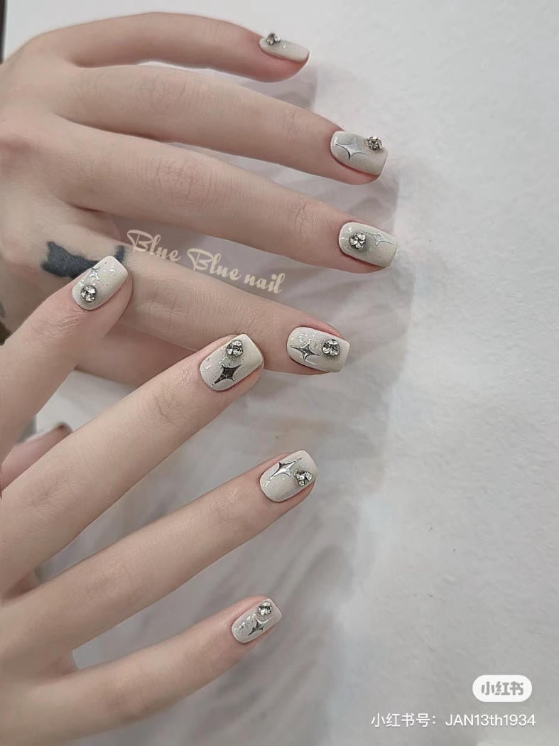 Bàn tay với móng tay được sơn màu trắng, trang trí đá và họa tiết tinh tế trên nền tường sáng.
