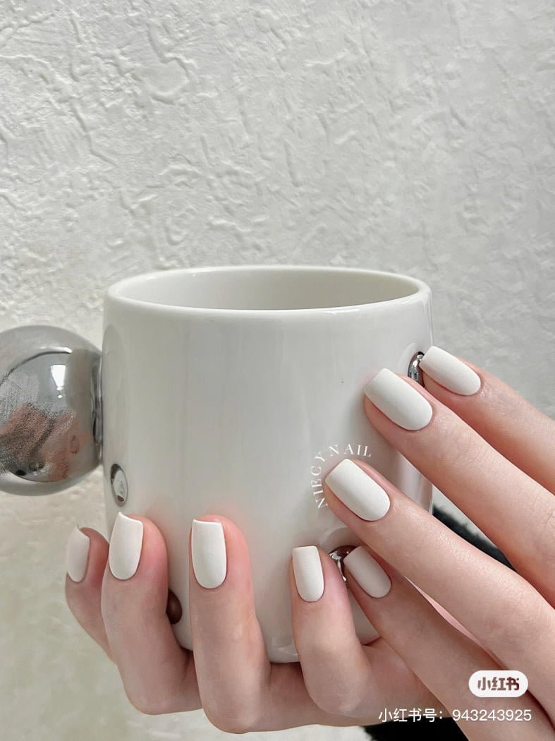 Bàn tay với móng tay sơn màu trắng, cầm một chiếc cốc trắng, nền tường màu trắng.