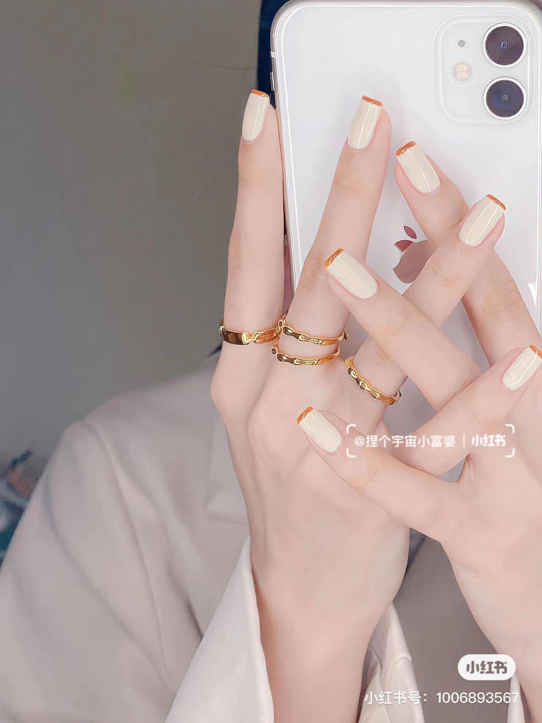 Người cầm điện thoại, móng tay sơn màu trắng với viền cam, đeo nhiều nhẫn vàng.